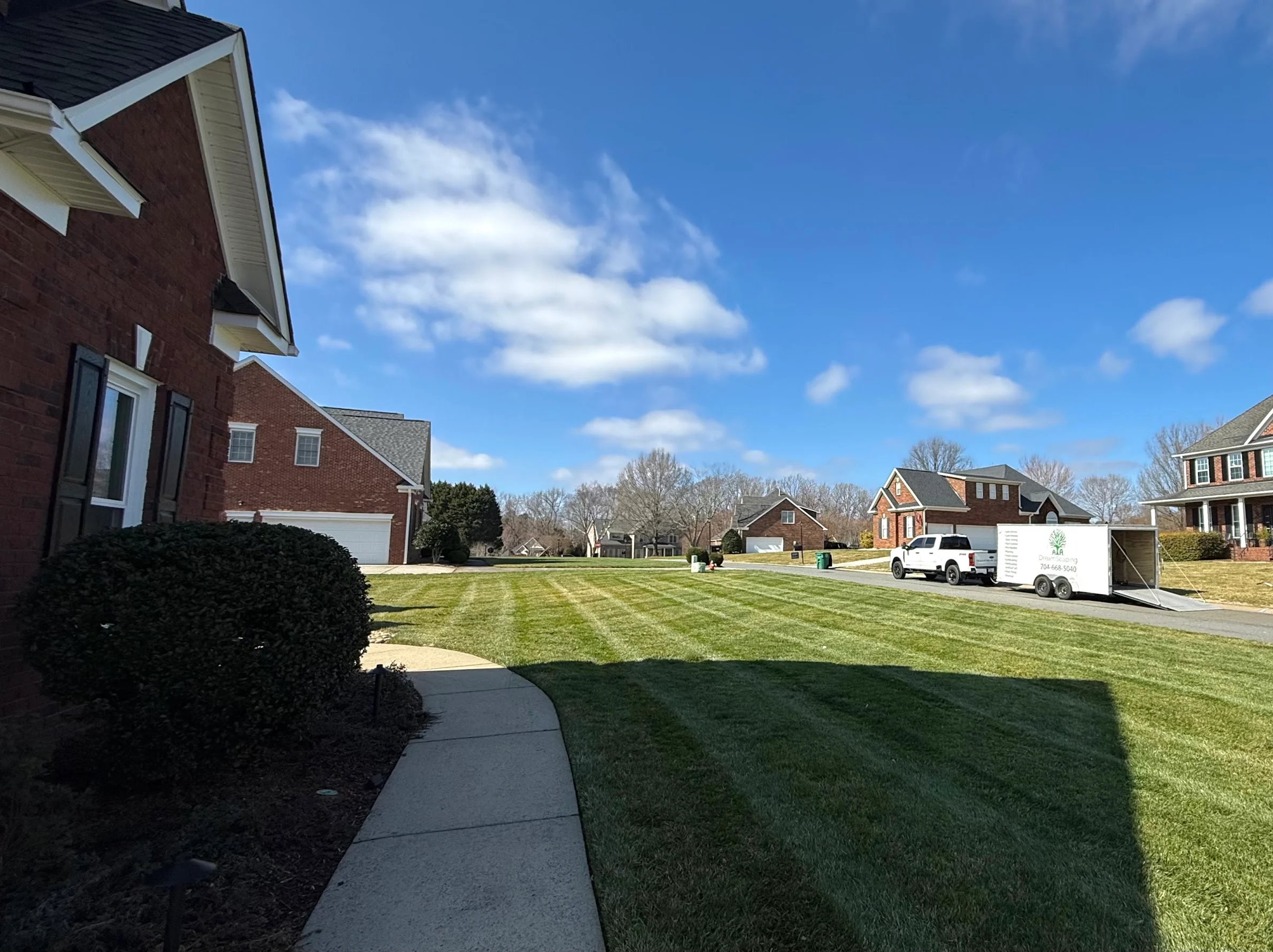 Striped lawn with manicured shrub beds and concrete walkway along brick home
