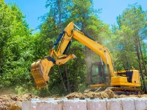 Yellow excavator with extended arm digging and moving soil on cleared residential lot with trees